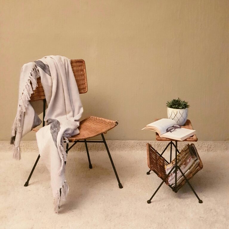 A cozy reading nook with a wicker chair, blanket, and small table featuring a book and plant.