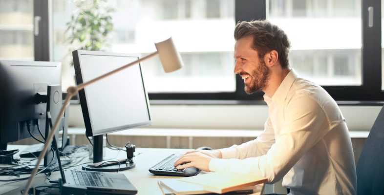 A man smiling while working at an office desk with a computer and natural daylight streaming in through large windows.
